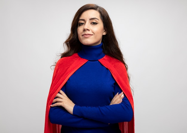 Confident caucasian superhero girl with red cape stands with crossed arms isolated on white wall with copy space