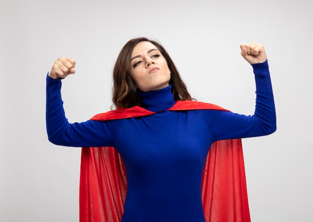 Confident caucasian superhero girl with red cape tenses biceps and looks at side on white