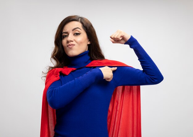 Confident caucasian superhero girl with red cape tenses and points at biceps on white