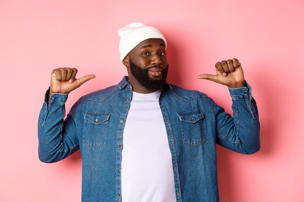 Confident and cheeky Black man pointing at himself, staring at camera self-assured, show-off, standing over pink background.