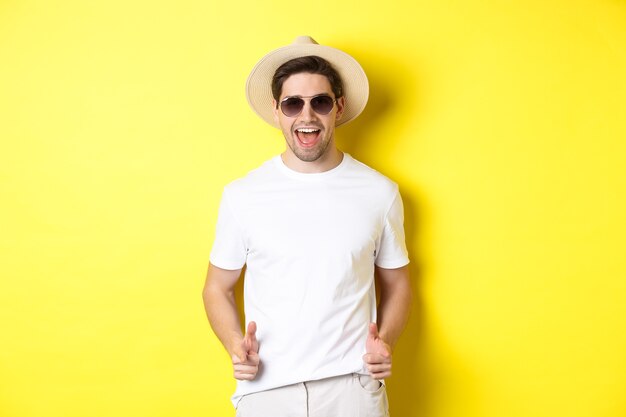 休暇中の自信を持って生意気な男があなたとイチャイチャして、カメラに人差し指を向けてウインクし、サングラス、黄色の背景の夏の帽子をかぶっています。