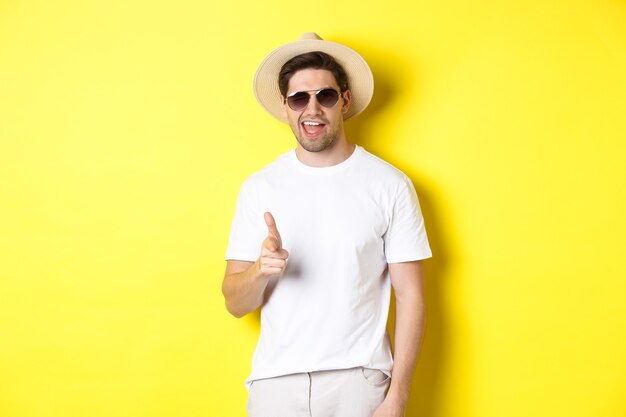 Confident and cheeky guy on vacation flirting with you, pointing finger at camera and winking, wearing summer hat with sunglasses, yellow background