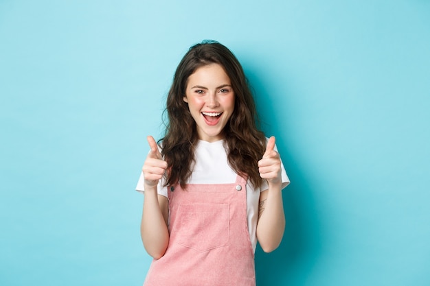 Confident and cheerful brunette girl say yes, pointing fingers at camera and looking sassy, praising you, inviting to event, standing over blue background