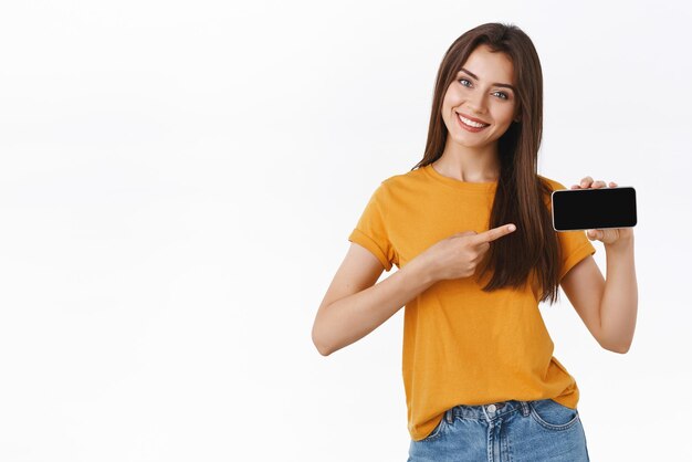 新しいアプリケーションアプリの機能やゲームの笑顔を喜んで紹介するようにスマートフォンを水平に向けたモバイル画面を保持している黄色のTシャツで自信を持って陽気なゴージャスな若い現代女性