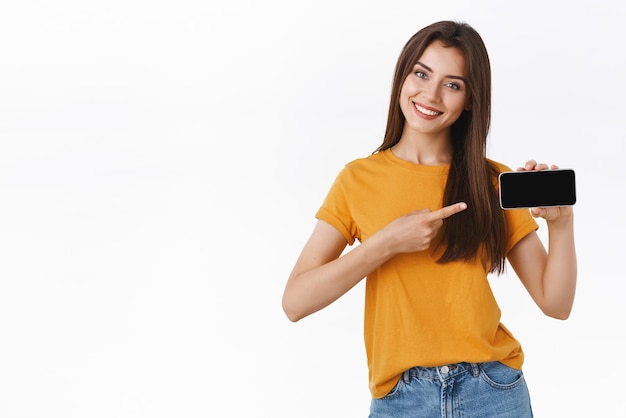 Уверенная веселая великолепная молодая современная женщина в желтой футболке держит смартфон горизонтально, указывая на мобильный экран, представляя новую функцию приложения или игру, улыбаясь в восторге