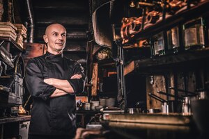 Confident chef wearing uniform posing with his arms crossed and looking away in a restaurant kitchen.