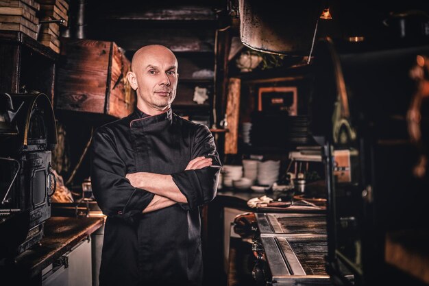 Confident chef wearing uniform posing with his arms crossed and looking at a camera in a restaurant kitchen.