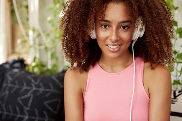 Уверенная смуглая привлекательная афроамериканка в розовой повседневной футболке слушает онлайн песни в наушниках, наслаждается любимой композицией, отдыхает дома. Люди, технологии, образ жизни