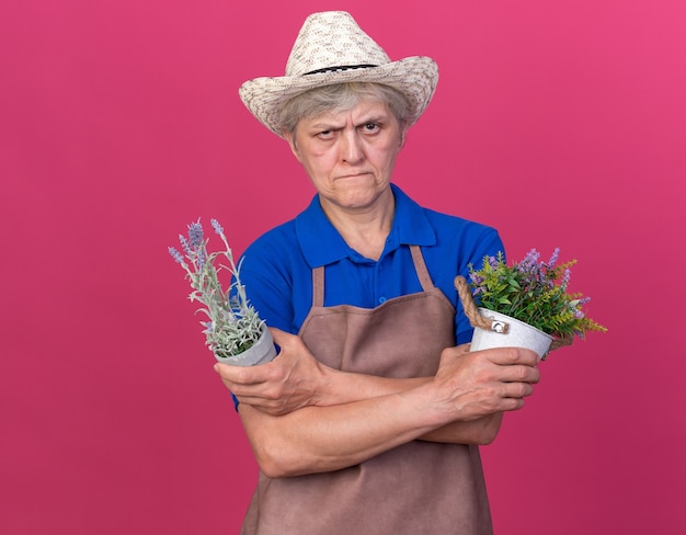 Confident elderly female gardener wearing gardening hat crossing arms holding flowerpots isolated on pink wall with copy space