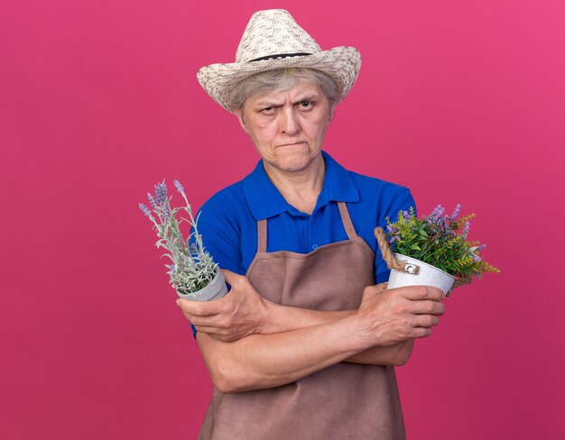 Confident elderly female gardener wearing gardening hat crossing arms holding flowerpots isolated on pink wall with copy space