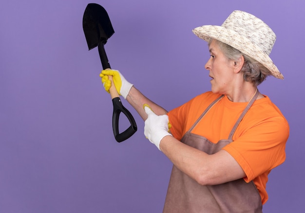 Free photo confident elderly female gardener wearing gardening hat and gloves holds and points at spade looking at side on purple