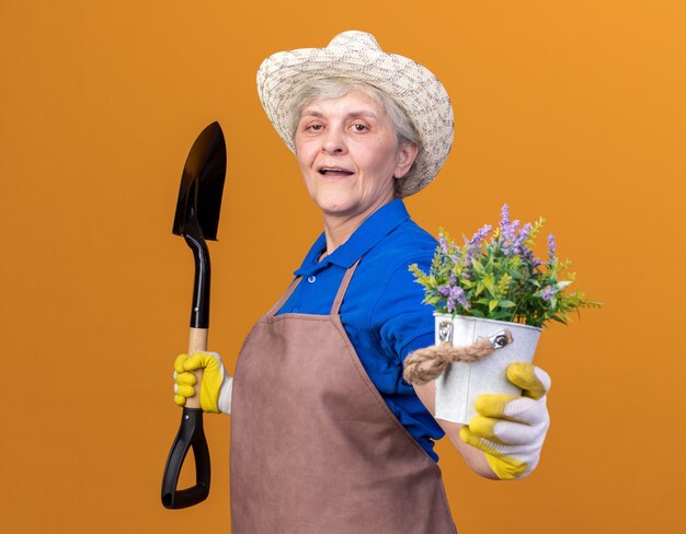 ガーデニングの帽子と手袋を身に着けている自信を持って年配の女性の庭師は、スペードと植木鉢を持って横に立っています