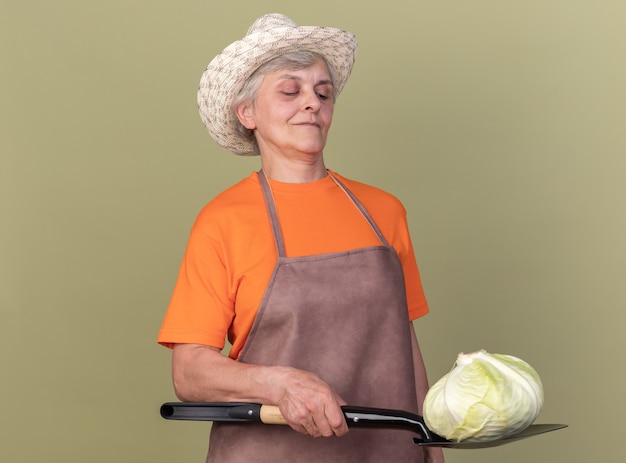 Бесплатное фото Уверенная пожилая женщина-садовник в садовой шляпе держит капусту на лопате