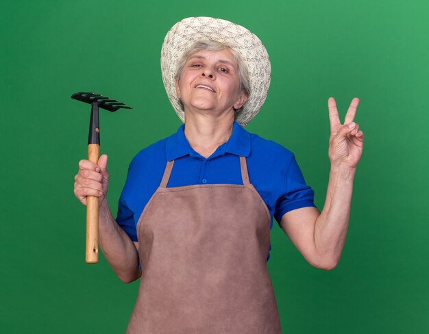 Уверенная пожилая женщина-садовник в садовой шляпе держит грабли и жестикулирует знак победы