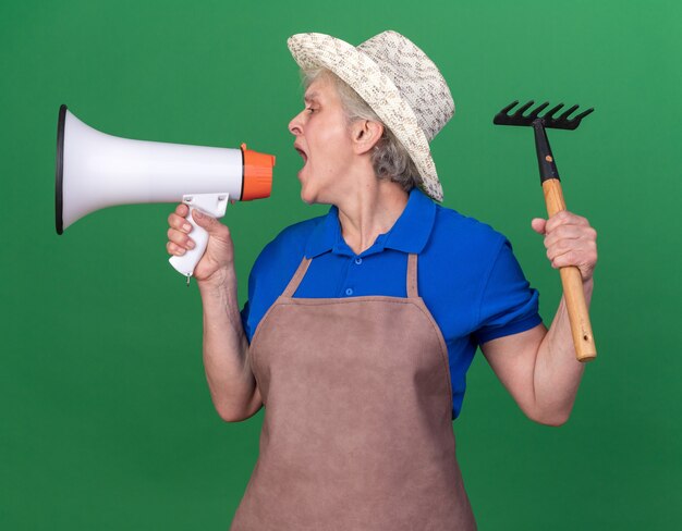 Confident elderly female gardener wearing gardening hat holding rake and shouting into loud speaker looking at side isolated on green wall with copy space