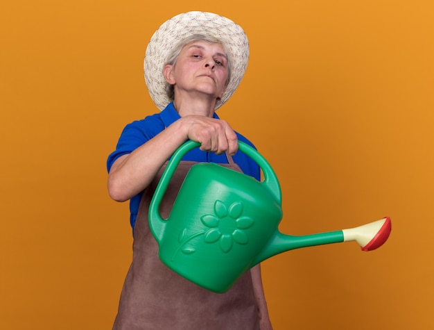 Confident elderly female gardener wearing gardening hat holding watering can