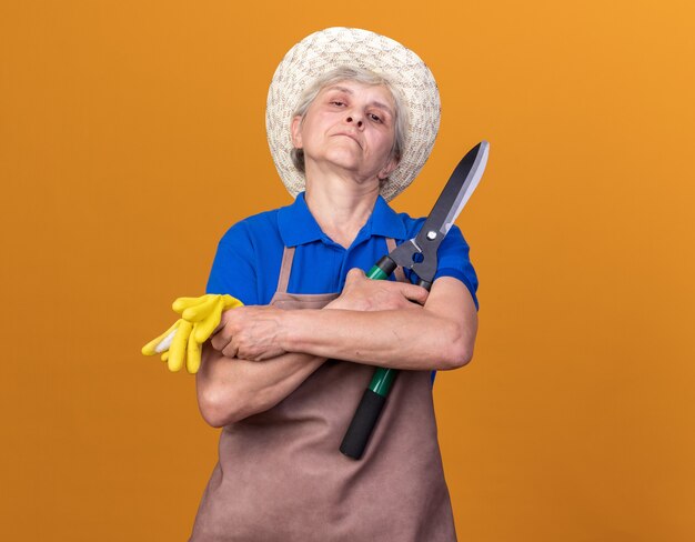 Confident elderly female gardener wearing gardening hat standing with crossed arms holding gardening scissors and gloves