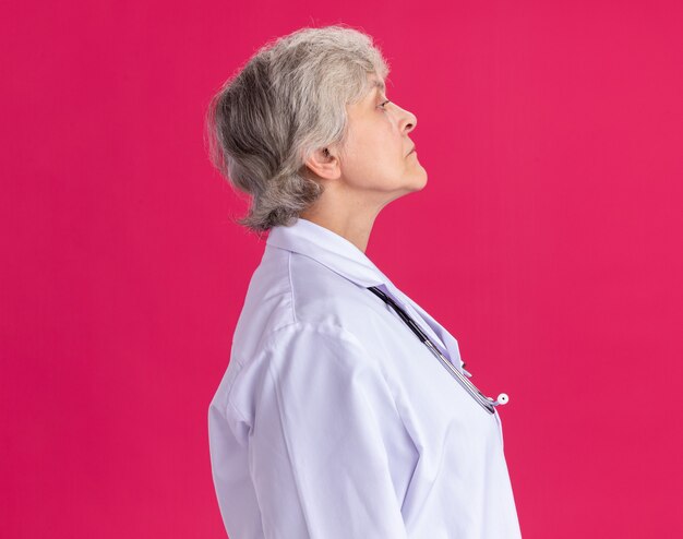 Confident elderly woman in doctor uniform with stethoscope standing sideways isolated on pink wall with copy space