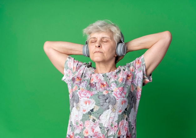 Free photo confident elderly woman on headphones stands holding hands behind head with closed eyes isolated on green wall