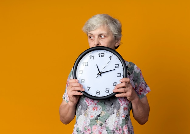 Уверенная пожилая женщина держит часы, глядя на сторону, изолированную на оранжевой стене