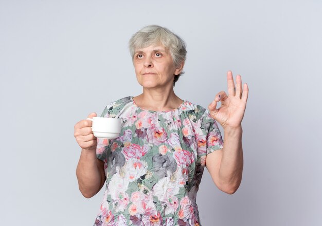 Уверенная в себе пожилая женщина держит чашку и жестикулирует знаком рукой, изолированной на белой стене