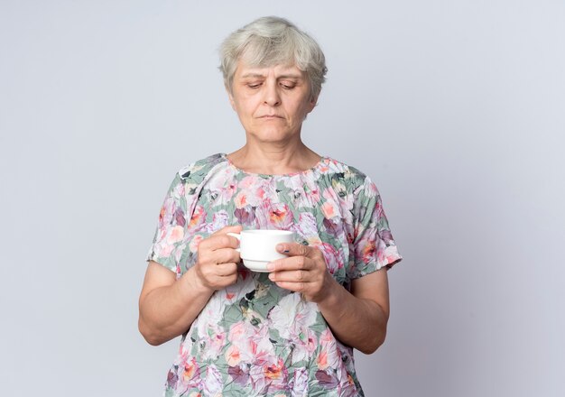 Confident elderly woman holds and looks at cup isolated on white wall