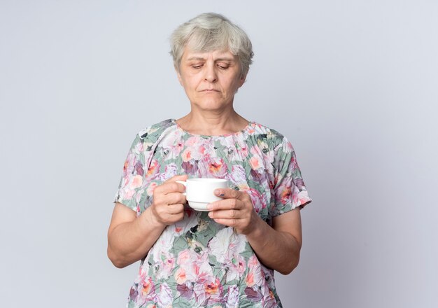 自信を持って年配の女性が白い壁に隔離されたカップを保持し、見ています