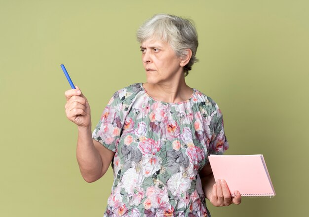 Confident elderly woman holds notebook and pen looking at side isolated on olive green wall