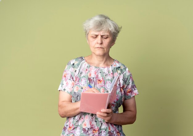 自信を持って年配の女性は、オリーブグリーンの壁に隔離された何かを書き留めるノートとペンを保持します。