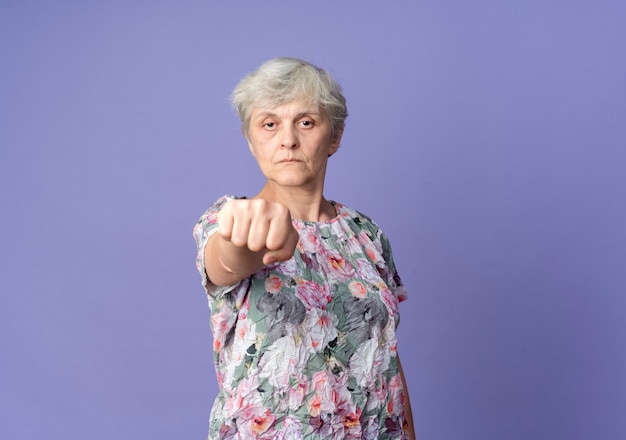 Confident elderly woman holds out fist isolated on purple wall