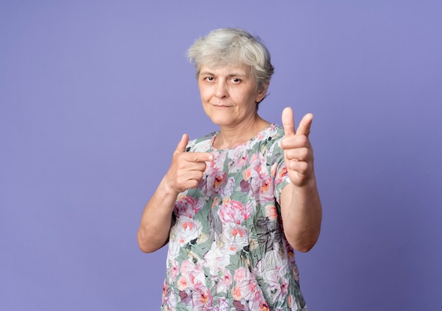 Confident elderly woman points with two hands isolated on purple wall