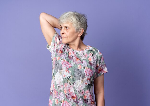 Confident elderly woman puts hand on head behind looking at side isolated on purple wall