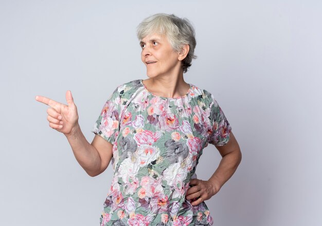 Confident elderly woman puts hand on waist looking and pointing at side isolated on white wall