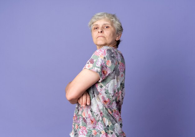 Confident elderly woman stands sideways looking isolated on purple wall