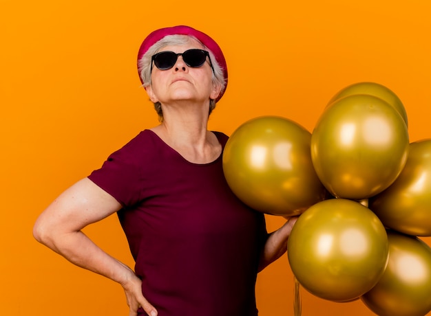 Уверенная в себе пожилая женщина в солнцезащитных очках в шляпе для вечеринки стоит с гелиевыми шарами, изолированными на оранжевой стене с копией пространства