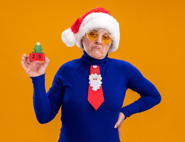 Confident elderly woman in sun glasses with santa hat and santa tie holding christmas tree ornament isolated on orange background with copy space
