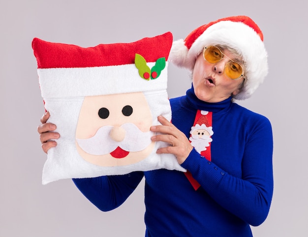 Free photo confident elderly woman in sun glasses with santa hat and santa tie holding santa pillow isolated on white background with copy space