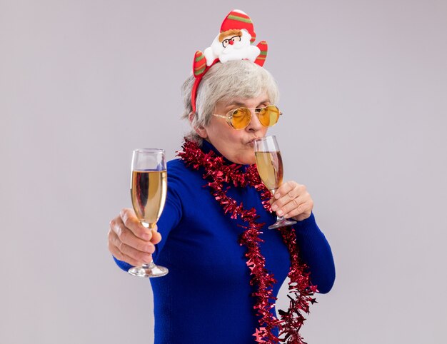 Confident elderly woman in sun glasses with santa headband and garland around neck holds and drinks glasses of champagne 