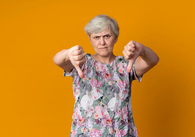 Confident elderly woman thumbs down with two hands isolated on orange wall