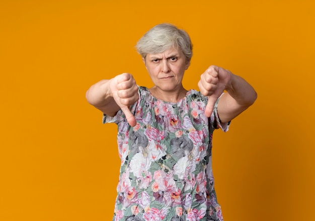 Уверенная пожилая женщина показывает палец вниз двумя руками, изолированными на оранжевой стене