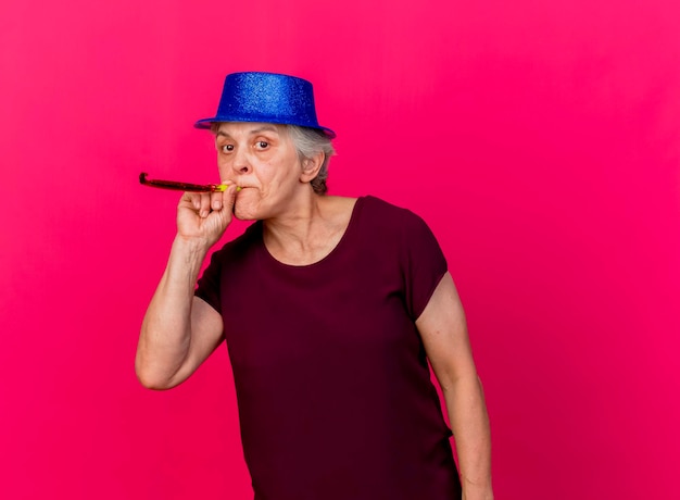 Confident elderly woman wearing party hat blowing whistle looking at camera on pink