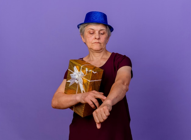 Confident elderly woman wearing party hat holds gift box looking at arm isolated on purple wall with copy space