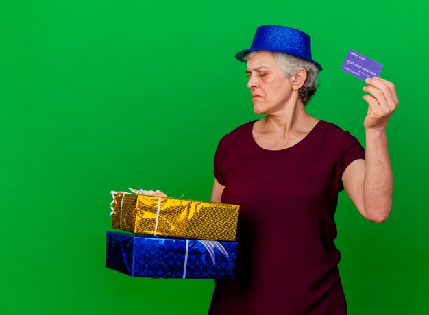 Confident elderly woman wearing party hat holds gift boxes and credit card on green