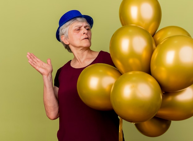 Confident elderly woman wearing party hat looks at helium balloons raising hand isolated on olive green wall with copy space
