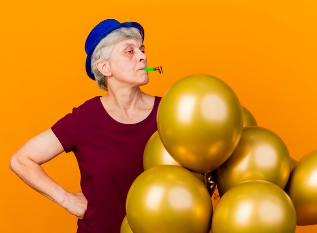 Confident elderly woman wearing party hat stands with helium balloons blowing whistle isolated on orange wall with copy space