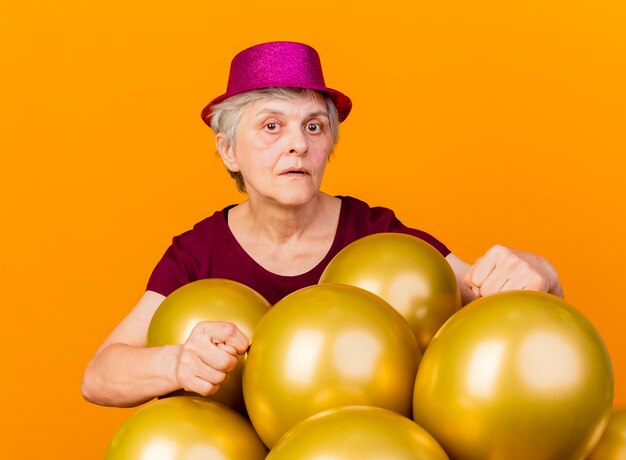 Confident elderly woman wearing party hat stands with helium balloons gesturing bathroom hand sign isolated on orange wall