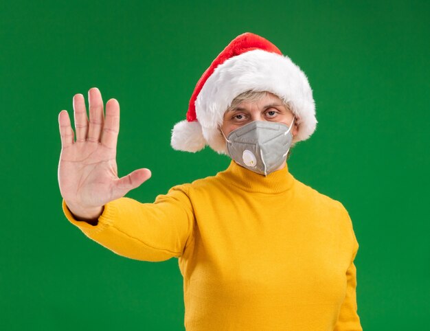 Confident elderly woman with santa hat wearing medical mask gesturing stop sign isolated on green background with copy space