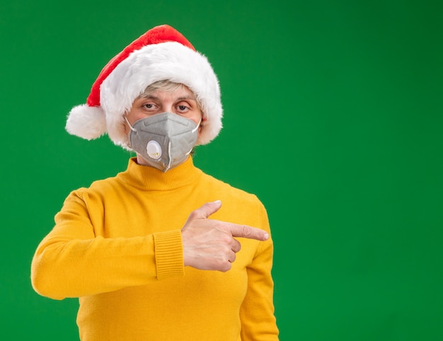 Confident elderly woman with santa hat wearing medical mask pointing at side isolated on green background with copy space