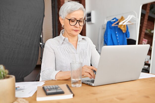 Уверенная в себе опытная зрелая деловая женщина в стильных очках печатает письма своим клиентам и деловым партнерам, используя высокоскоростное беспроводное подключение к Интернету на ноутбуке.
