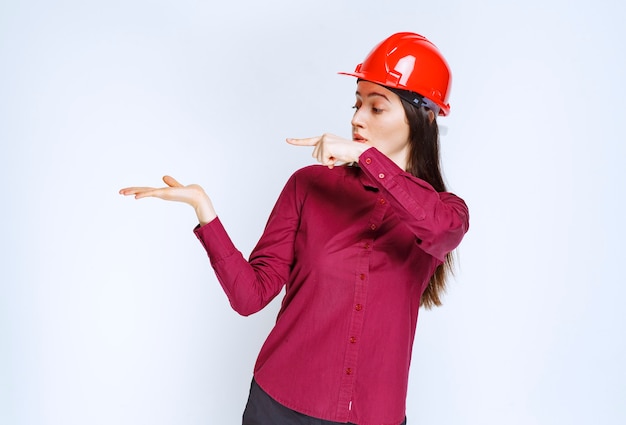 Confident female architect in red hard helmet standing and pointing. 